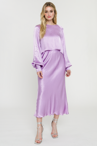 Model wearing a purple, satin, midi, skirt. She is standing in front of a white backdrop. The model is wearing a matching top with the skirt. 