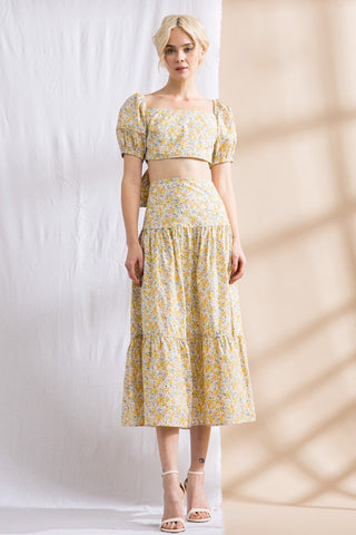 Model standing in front of white and tan backdrop. Wearing a flower patterned, two-piece, skirt set. Sleeves are puffy.