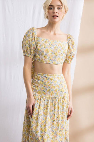 Model standing in front of white and tan backdrop. Wearing a flower patterned, two-piece, skirt set. Sleeves are puffy.
