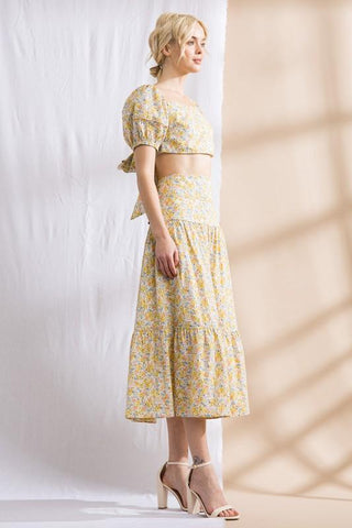 Model standing in front of white and tan backdrop. Wearing a flower patterned, two-piece, skirt set. Sleeves are puffy.Model standing in front of white and tan backdrop. Wearing a flower patterned, two-piece, skirt set. Sleeves are puffy. Model is facing to the side.