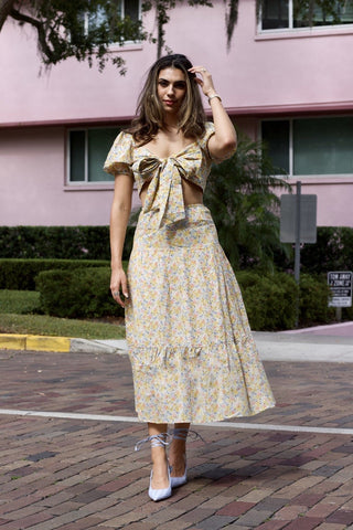 Model standing in front of pink building. Wearing a flower patterned, two-piece, skirt set. Sleeves are puffy and bow is tied in the front. Model is walking towards the camera.