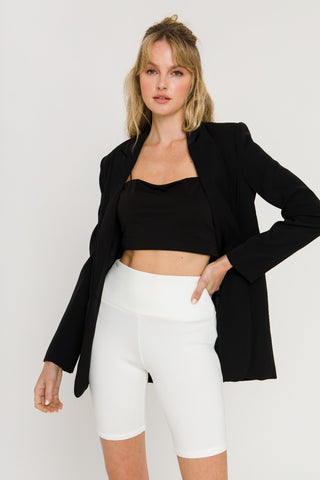 Model wearing white, mid-thigh, biker shorts with black crop top and black blazer. She is standing in front of a white backdrop.