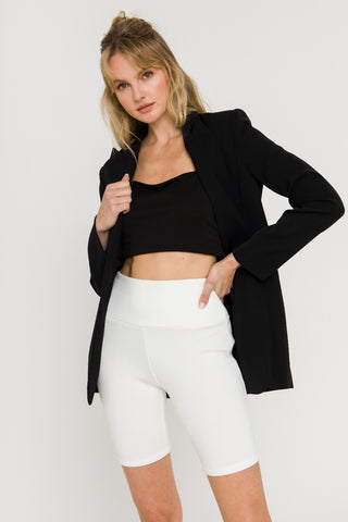 Model wearing white, mid-thigh, biker shorts with black crop top and black blazer. She is standing in front of a white backdrop. She is grabbing one side of her blazer collar with her hand.