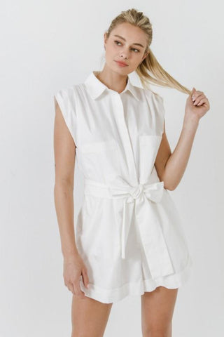 Model wearing white, collared, cotton, belted romper. She is standing in front of a white backdrop and is looking into the camera. The front of the romper is showing. The model is playing with her hair. 