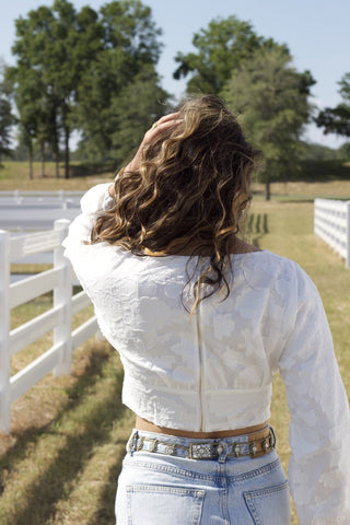 Model standing in front of a fence and grass. She is wearing a white, flower textured, long sleeve top and denim. The photos is from behind to show the details of the back and the gold zipper on the back of the shirt.