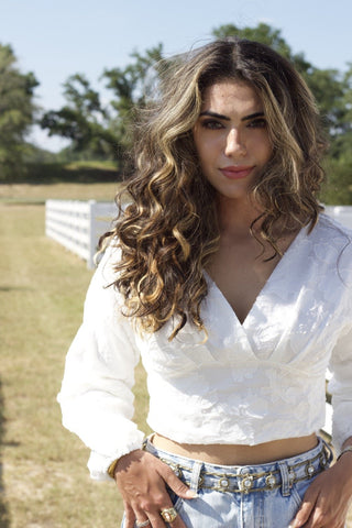 Model standing in front of a fence and grass. She is wearing a white, flower textured, long sleeve top and denim. 