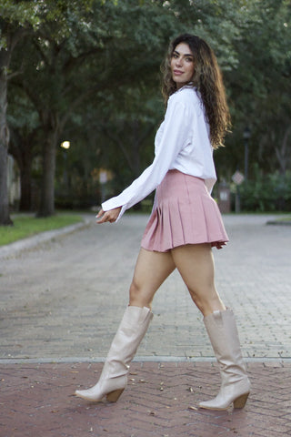 Girl walking across street in blush pleated tennis skirt and white button up. Background is road and trees.