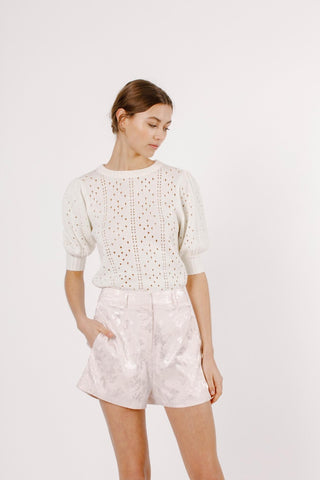 Model wearing pink, satin, jacquard shorts with short sleeve sweater top. Model is standing in front of white back drop and looking down.