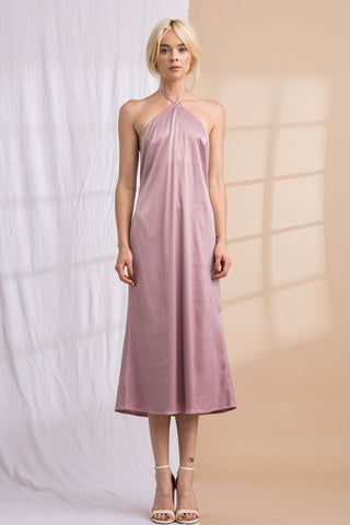 Model wearing a purple, mauve, satin, halter, midi dress. Model standing in front of a backdrop that is half white and half tan. 